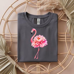 May include: A dark gray t-shirt featuring a pink flamingo with a peony flower body. The flamingo has a black beak and pink legs. The shirt is folded and placed on a woven wicker chair.