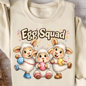 Puede incluir: Sudadera color crema con el texto "Egg Squad" sobre tres ovejas de dibujos animados con orejas de conejo y huevos de Pascua. Las ovejas están en varias poses, con acentos coloridos.