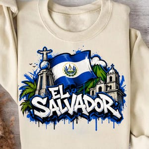 May include: Cream-colored sweatshirt featuring a graphic design of El Salvador. The design includes the Salvadoran flag, a monument, a church, and the text "EL SALVADOR" in a graffiti style with blue accents.