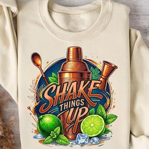 Könnte beinhalten: Ein cremefarbenes Sweatshirt mit einer Grafik aus einem kupferfarbenen Cocktailshaker, einem Rührlöffel und einem Messbecher. Das Design zeigt leuchtend grüne Limetten, Minzblätter, Eiswürfel und den Text SHAKE THINGS UP in orangefarbenen Buchstaben.