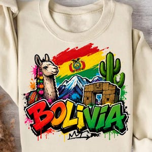 Puede incluir: Una sudadera color crema con un estampado gráfico de una llama con borlas coloridas, la bandera de Bolivia, montañas nevadas, un cactus verde y un arco de piedra. La palabra BOLIVIA está impresa en letras grandes, negritas y estilo grafiti.