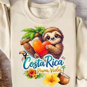 Könnte beinhalten: Cremefarbenes Sweatshirt mit einem Cartoon-Faultier, das ein Herz hält, mit dem Text "Costa Rica Pura Vida". Das Design umfasst einen Tukan, Palmblätter und tropische Blumen. Eine Kokosnuss ist ebenfalls abgebildet.