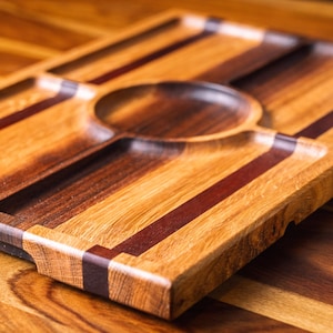 Puede incluir: Bandeja de madera hecha a mano con un cuenco central circular y cuatro compartimentos rectangulares. La bandeja presenta una rica paleta de colores cálidos de marrones claros y oscuros con acentos rojo oscuro. La veta de la madera es visible.