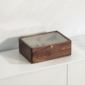 May include: A wooden jewelry box with a clear glass top. The box is rectangular with a dark brown finish and a small gold-colored clasp. The interior is divided into compartments with a light gray lining.