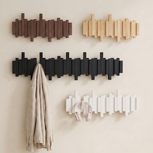 May include: A display of wooden wall-mounted coat racks in various colors: dark brown, natural wood, black, and white. A beige shirt hangs on the black rack, and pink headphones rest on the white rack. The racks feature vertical wooden slats.