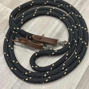 May include: Black and cream-coloured rope dog lead with leather accents and a silver-coloured clasp. The lead is coiled and features a repeating pattern of cream-coloured squares. The leather accents are brown.