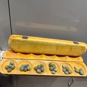 May include: A wooden mancala board with a hinged lid, featuring six circular depressions filled with small, grey-green stones. The board is made of light-coloured wood with carved details, suggesting a traditional game.