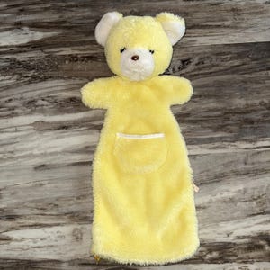 May include: A yellow plush bear hand puppet with white accents on the ears and a white pocket. The bear has a brown nose and black eyes. The puppet is made of soft, fuzzy material.