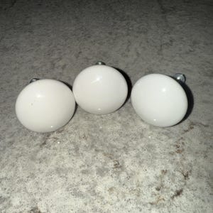 May include: Three white, round cabinet knobs with silver hardware. The knobs are evenly spaced and appear to be made of ceramic or porcelain. They are set against a speckled gray surface.