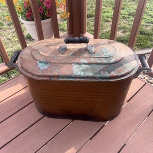 May include: An antique copper oval-shaped cooking pot with a lid. The pot has a dark handle on top and two side handles. The copper has a patina with green and brown hues. The pot is sitting on a wooden deck.