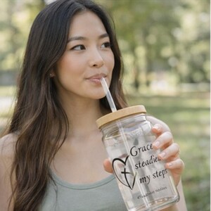 May include: A clear glass tumbler with a bamboo lid and straw. The tumbler has the text "Grace steadies my steps" with a heart and cross design. The person is drinking from the tumbler.