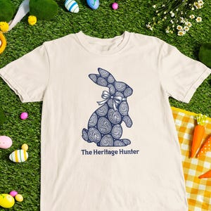 May include: A cream-coloured t-shirt with a navy blue paisley bunny design and the text "The Heritage Hunter". The shirt is displayed on a green surface with Easter decorations, including eggs, carrots, and a stuffed bunny.