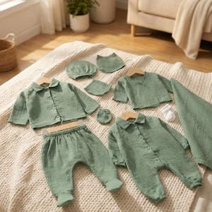 Könnte beinhalten: Eine Kollektion von babykleidung in Salbeigrün, darunter ein Hemd, eine Hose, ein Strampler, eine Mütze und Fäustlinge. Die Artikel sind aus einem weichen, strukturierten Stoff gefertigt und auf einer cremefarbenen Decke präsentiert.