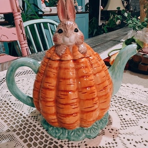 以下が含まれることがあります： ウサギが上に乗った、ニンジン型のセラミックティーポット。 ティーポットはオレンジ色で、緑と青のアクセントが付いています。 ウサギは茶色の毛皮とピンクの耳をしています。 ティーポットは白いレースのテーブルクロスの上に置かれています。