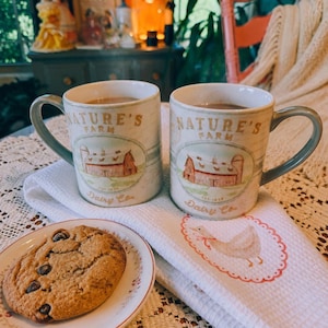 May include: Two ceramic mugs with the text "NATURE'S FARM Dairy Co." and a barn illustration, filled with a brown liquid, sit on a white towel. A chocolate chip cookie rests on a small plate. The scene evokes a cozy, rustic aesthetic.