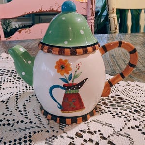 May include: A decorative ceramic teapot with a white body, green lid with white polka dots, and a blue knob. The teapot features a floral design and a brown and orange striped handle. The spout is green with white polka dots.
