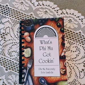 May include: A spiral-bound cookbook titled "What's Phi Mu Got Cookin'" with a cover featuring illustrations of food items. The cookbook is from the Phi Mu Fraternity, Zeta Lambda. The book is on a white lace tablecloth.