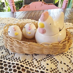 Puede incluir: Un juego de cocina que incluye un recipiente de cerámica blanca con forma de gallina con cresta rosa y pico amarillo, junto con dos saleros y pimenteros con forma de pollito, en una cesta de mimbre.
