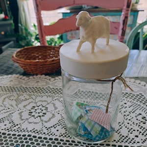 May include: A clear glass jar with a white lid, topped with a small, off-white sheep figurine. Inside the jar are colorful fabric squares. The jar is tied with twine and sits on a white lace tablecloth.