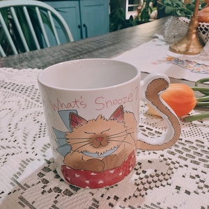 May include: White ceramic mug with a whimsical illustration of a sleeping cat. The mug features a curled tail handle and the text "What's Snooze?" in pink. The cat is light brown and resting on a red and white polka dot pillow.
