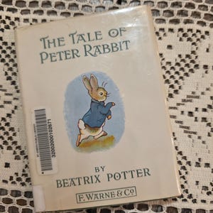 Peut inclure: Un livre de poche intitulé "The Tale of Peter Rabbit" de Beatrix Potter. La couverture présente une illustration de Pierre Lapin en veste bleue. Le titre est en vert, et l'éditeur est F. Warne & Co. Une étiquette de bibliothèque est collée sur le dos.