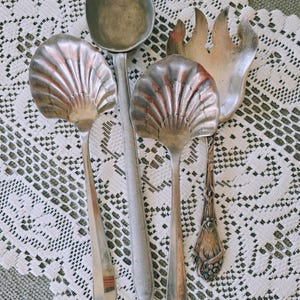 May include: A collection of antique silver serving utensils, including two shell-shaped spoons, a large ladle, and a serving fork. The utensils are arranged on a white lace tablecloth, showcasing their intricate designs and aged patina. The spoons have a fan-like design.