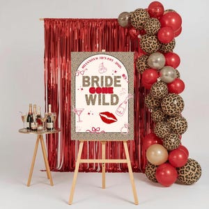 Peut inclure: Un chevalet en bois présente une affiche avec le texte "Bride Gone Wild" en rouge et imprimé léopard. L'affiche est ornée de rouge à lèvres, de champagne et d'illustrations de cocktails. Des ballons rouges et imprimés léopard et un rideau à franges rouge sont en arrière-plan.
