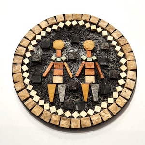Pode incluir: Obra de arte em mosaico redondo com duas figuras estilizadas. As figuras são compostas por várias telhas coloridas, incluindo laranja, bege e cinza. As figuras estão sobre um fundo escuro e texturizado, emolduradas por uma borda de telhas quadradas.