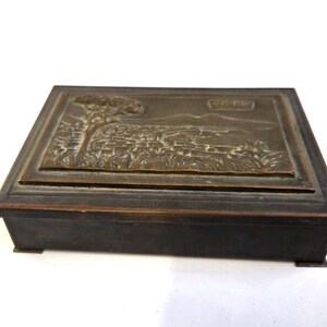 Puede incluir: Una caja rectangular de estilo antiguo con tapa color bronce. La tapa presenta un relieve detallado de un paisaje con un árbol y edificios. La base es de color marrón oscuro.