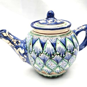 May include: A ceramic teapot with a lid, featuring a detailed blue, green, and white floral pattern. The spout and handle are also blue, with white and green accents. The teapot has a bulbous body and a rounded lid.
