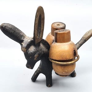 Puede incluir: Un juego de salero y pimentero con forma de burro. El burro es de color marrón oscuro con orejas largas y una cara detallada. Los saleros y pimenteros son de color marrón claro, sujetos por un anillo de latón y tienen una tapa de madera con agujeros.