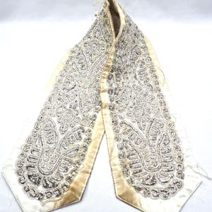 May include: An ivory-colored, decorative collar with intricate silver beading and a satin trim. The collar features an ornate, paisley-like design, with a cream-colored satin lining. The collar is likely vintage or antique.