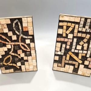 Pode incluir: Dois suportes de livros decorativos com designs de mosaicos. Cada suporte de livros tem uma forma retangular com a parte superior inclinada. Os mosaicos são em tons de bege, preto e castanho, dispostos em padrões abstratos.