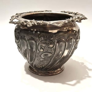 May include: An antique silver-toned bowl with detailed floral and scrollwork. The bowl has a decorative rim with floral accents and small handles. The body features vertical, curved panels with engraved designs. It sits on a small, circular base. A classic piece.