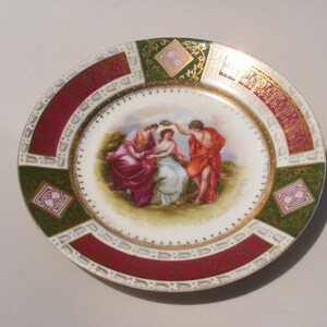 May include: A decorative porcelain plate with a central scene of figures in classical attire. The plate features a maroon and green border with gold accents and a detailed painted design. The plate is round and has a white background.