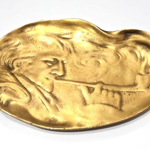 May include: A gold-colored, oval-shaped decorative tray. The tray features a relief of a person playing a wind instrument. The person's face and instrument are detailed in the metal. The tray has a smooth, reflective surface.