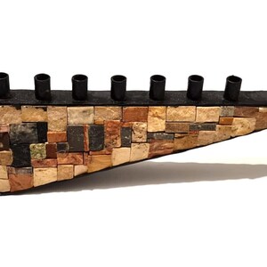 Pode incluir: Menorá artesanal com design de mosaico. A base é feita de pequenas pedras de formato irregular em tons de castanho, bege e cinza. Os castiçais são pretos e o braço curvo adiciona um toque decorativo.