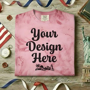 Puede incluir: Una camiseta rosa teñida con la frase "Your Design Here" en escritura negra. La camiseta está rodeada de decoraciones patrióticas, incluyendo una bandera americana, una brújula y una figura de la Estatua de la Libertad.