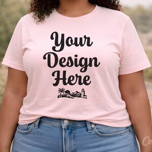 May include: A light pink t-shirt with the text "Your Design Here" in black script. Below the text is a graphic of a palm tree, mountains, and a lighthouse. The model is wearing blue jeans.