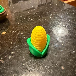 May include: A small, three-dimensional object shaped like an ear of corn with a bright yellow cob and textured kernels, partially encased in a vibrant green husk. The object has a smooth, matte finish and a rounded, upright, conical structure.