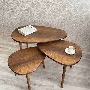 Puede incluir: Juego de tres mesas nido de madera con diseño de lágrima. Las mesas son de color marrón cálido y tienen patas delgadas y cónicas. Un libro descansa sobre la mesa superior, y una taza y platillo están en la mesa del medio.