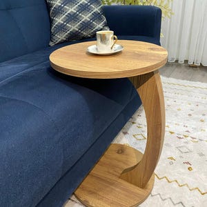 Puede incluir: Una mesa auxiliar de madera con tapa redonda y pata curva. La mesa es de madera marrón claro y está junto a un sofá azul marino. Una pequeña taza de café está sobre la mesa. La tapa de la mesa tiene unos 46 cm de diámetro.