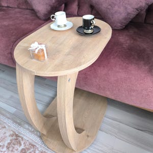 Pode incluir: Uma mesa lateral de madeira clara com um design único de pernas curvas. Duas xícaras de café pequenas estão sobre a mesa, uma branca e outra preta, cada uma com um pires. A mesa está em frente a um sofá malva com almofadas combinando.