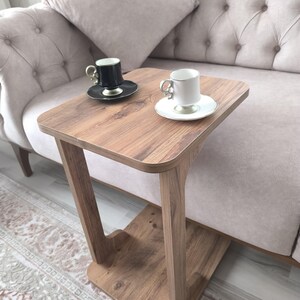 Op de afbeelding: Een kleine, houten bijzettafel met een vierkant blad en een uniek pootontwerp. Twee kleine koffiekopjes met schotels staan op de tafel. Eén kopje is zwart, de andere wit. De tafel staat naast een gecapitonneerde bank.