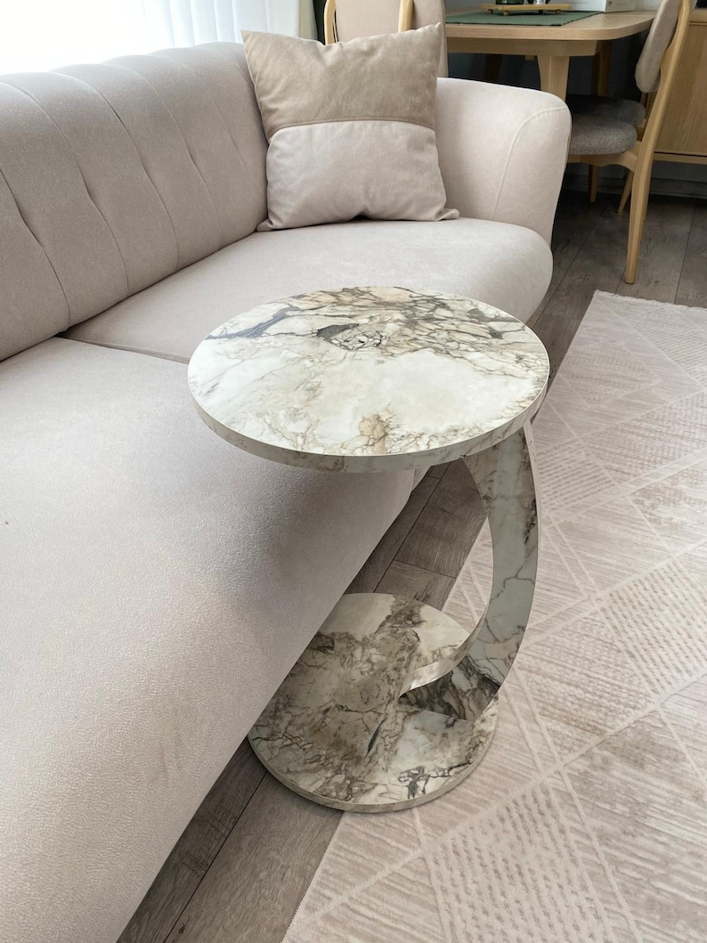 Op de afbeelding: Een ronde bijzettafel met een marmeren blad en een gebogen, marmerpatroon voet. De tafel staat naast een beige bank en een patroonkleed. Het tafelblad heeft een diameter van ongeveer 50 cm.
