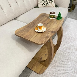 May include: A wooden side table with a warm wood grain finish. The table has a curved base and a rectangular top. A small teacup and saucer with orange accents sit on the table, along with a miniature house and a green Christmas tree.