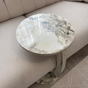 Op de afbeelding: Een ronde marmeren bijzettafel met een wit en grijs geaderd patroon. De tafel heeft &eacute;&eacute;n gebogen poot en staat naast een beige bank. De tafel heeft een diameter van ongeveer 50 cm.