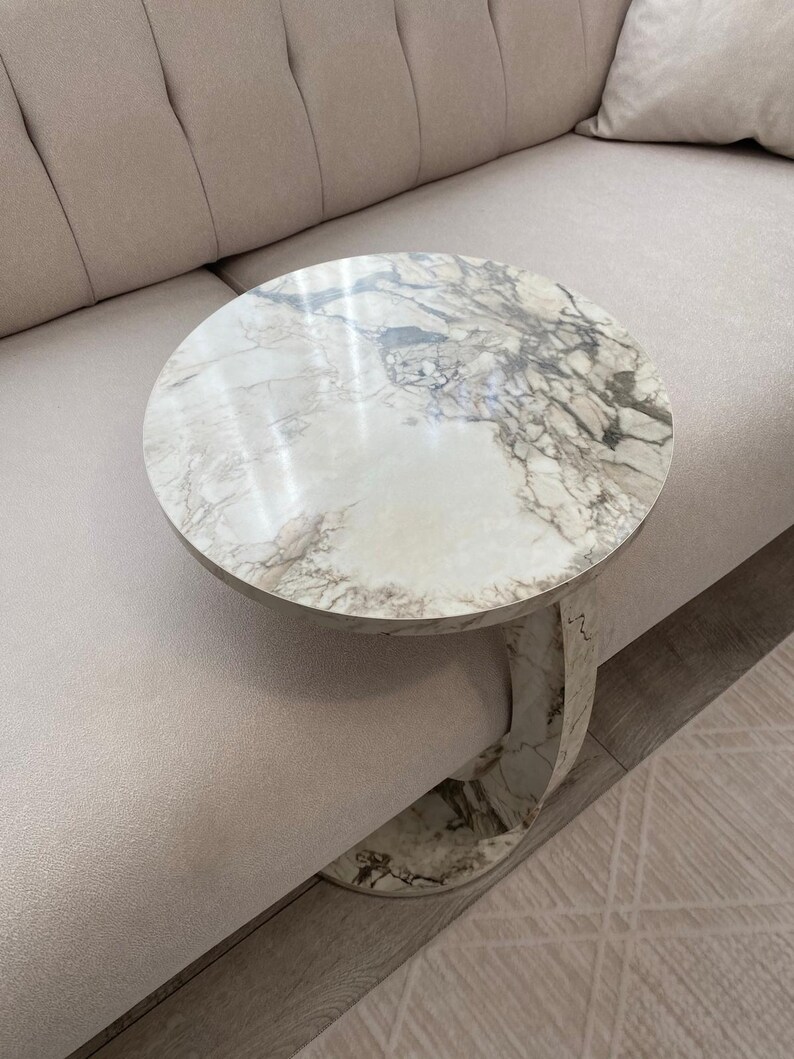 Op de afbeelding: Een ronde marmeren bijzettafel met een wit en grijs geaderd patroon. De tafel heeft &eacute;&eacute;n gebogen poot en staat naast een beige bank. De tafel heeft een diameter van ongeveer 50 cm.