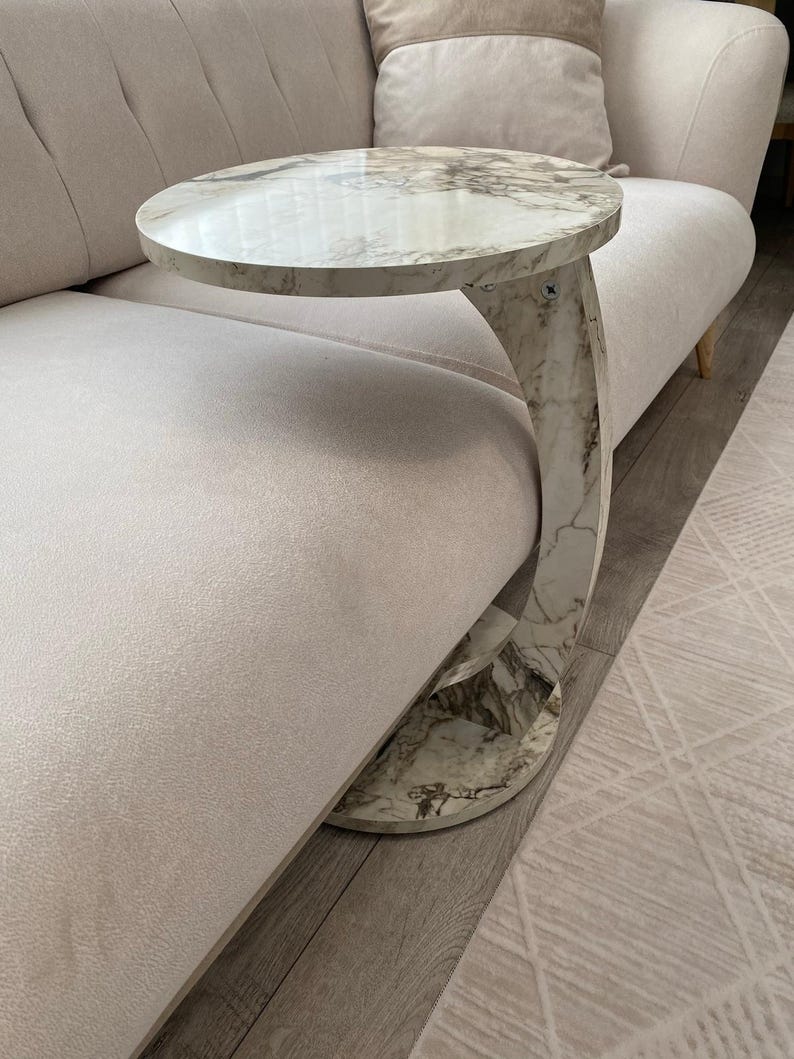 Op de afbeelding: Een bijzettafel met een marmeren blad en een gebogen poot. Het ronde tafelblad en de gebogen voet hebben een wit en grijs marmerpatroon. De tafel staat naast een beige bank en biedt een stijlvol accent.