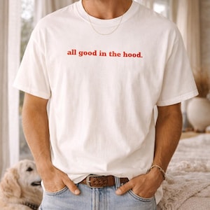 Puede incluir: Camiseta blanca con el texto rojo "all good in the hood". La camiseta se combina con vaqueros azules y un cinturón de cuero marrón. También se ven una cadena y una pulsera doradas. La persona está en el interior.